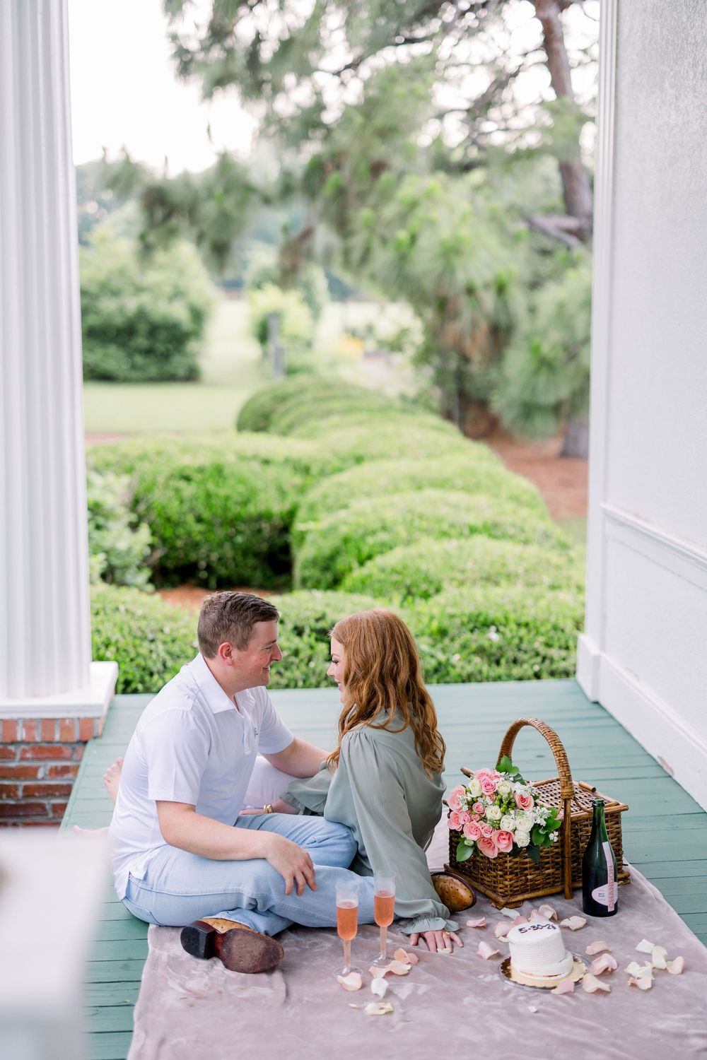 A couple enjoys a romantic picnic with wine and a wicker basket on a white porch with green trim and manicured gardens.