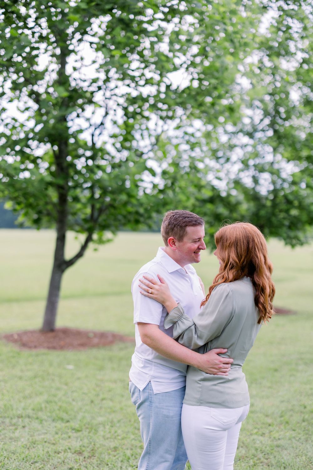 A couple embraces tenderly while standing beneath blooming trees in a grassy field.