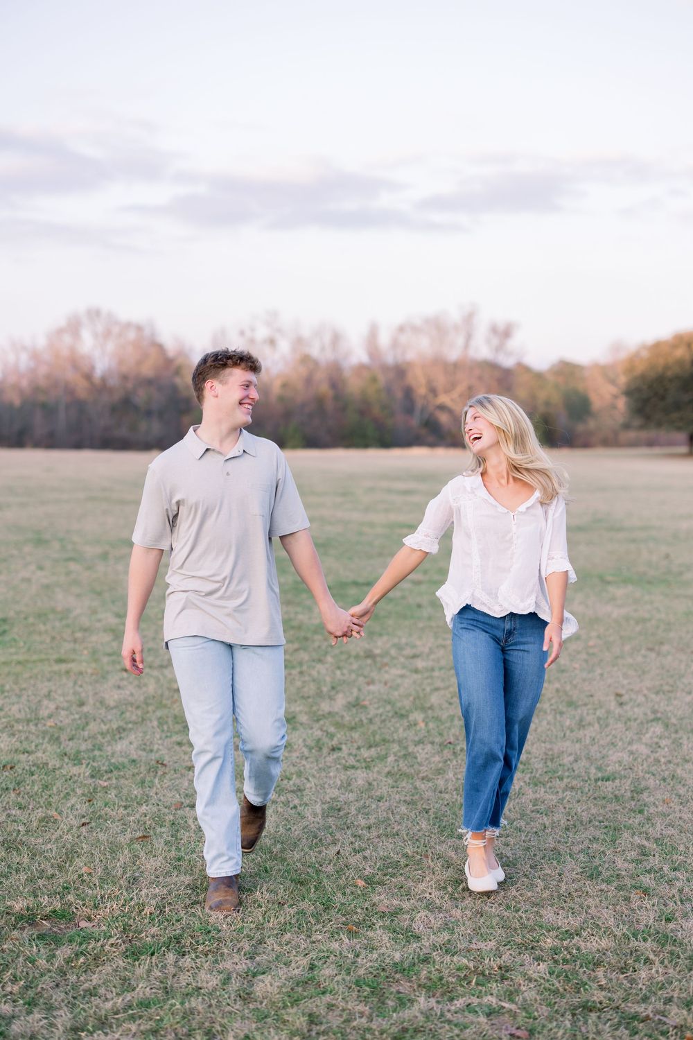 A couple walks hand in hand through an autumn field at sunset while laughing together.