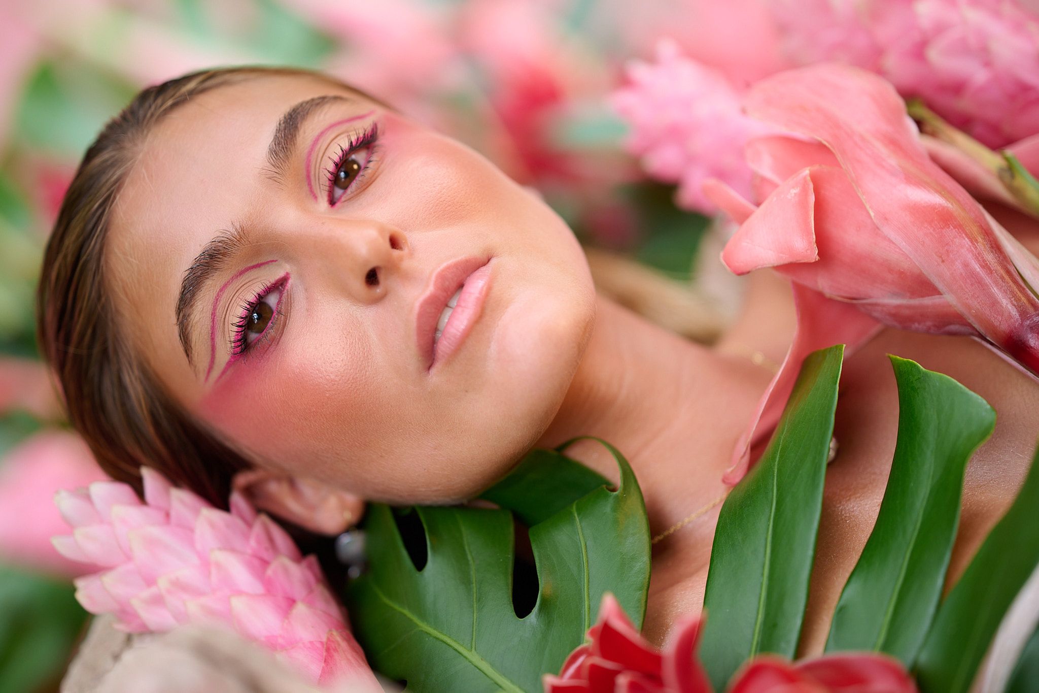 Soft pink flowers and tropical monstera leaves create a dreamy beauty portrait with natural lighting and rosy makeup.