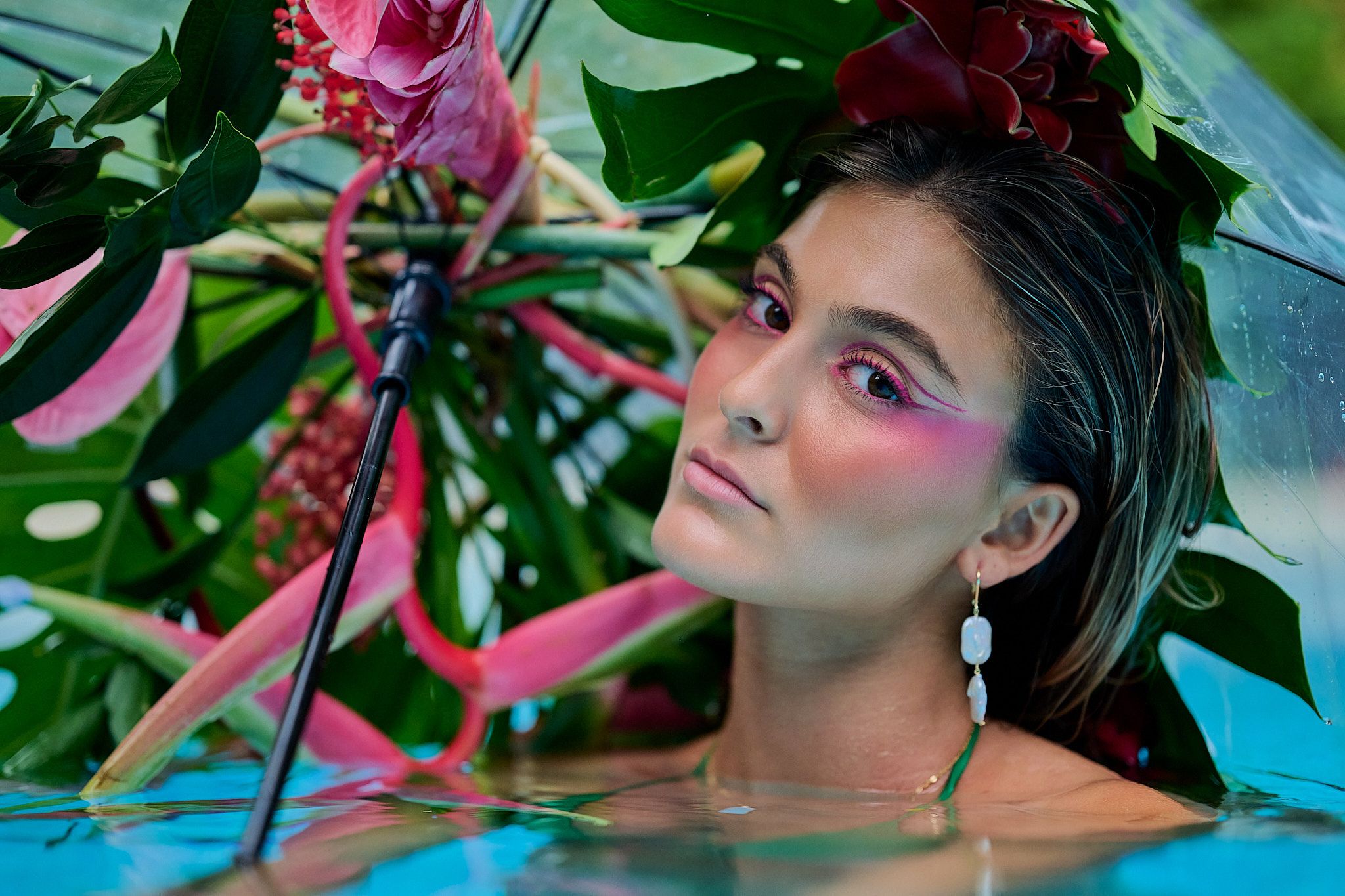Artistic beauty portrait with bright pink eye makeup and wet hair effect against turquoise water and green foliage.
