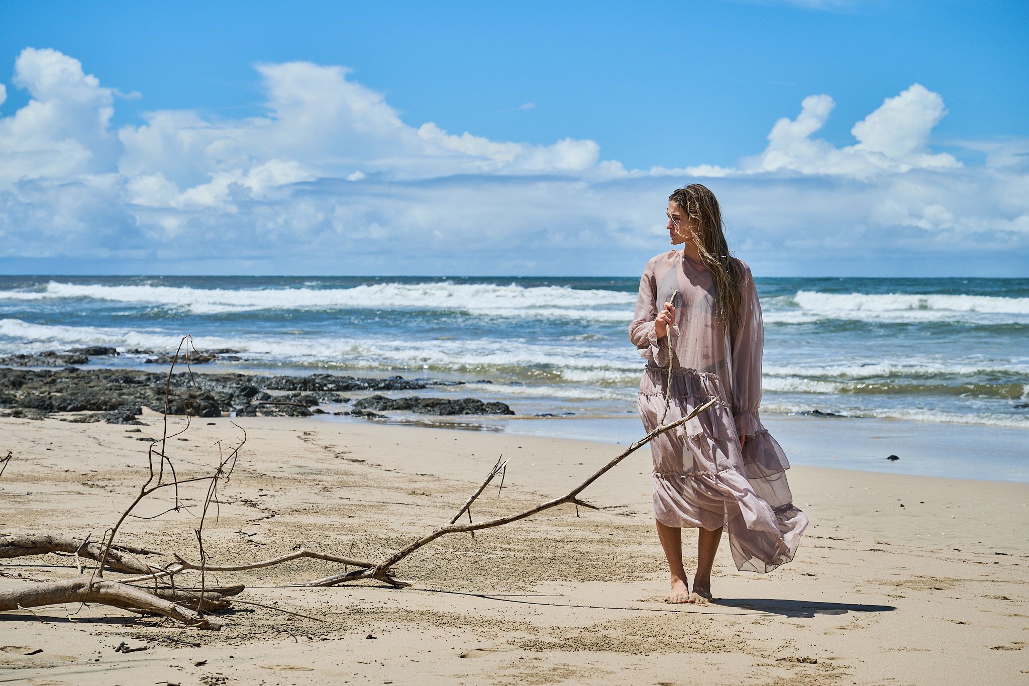 A person in a flowing dress walks along a scenic beach with crashing waves and blue skies in the background.