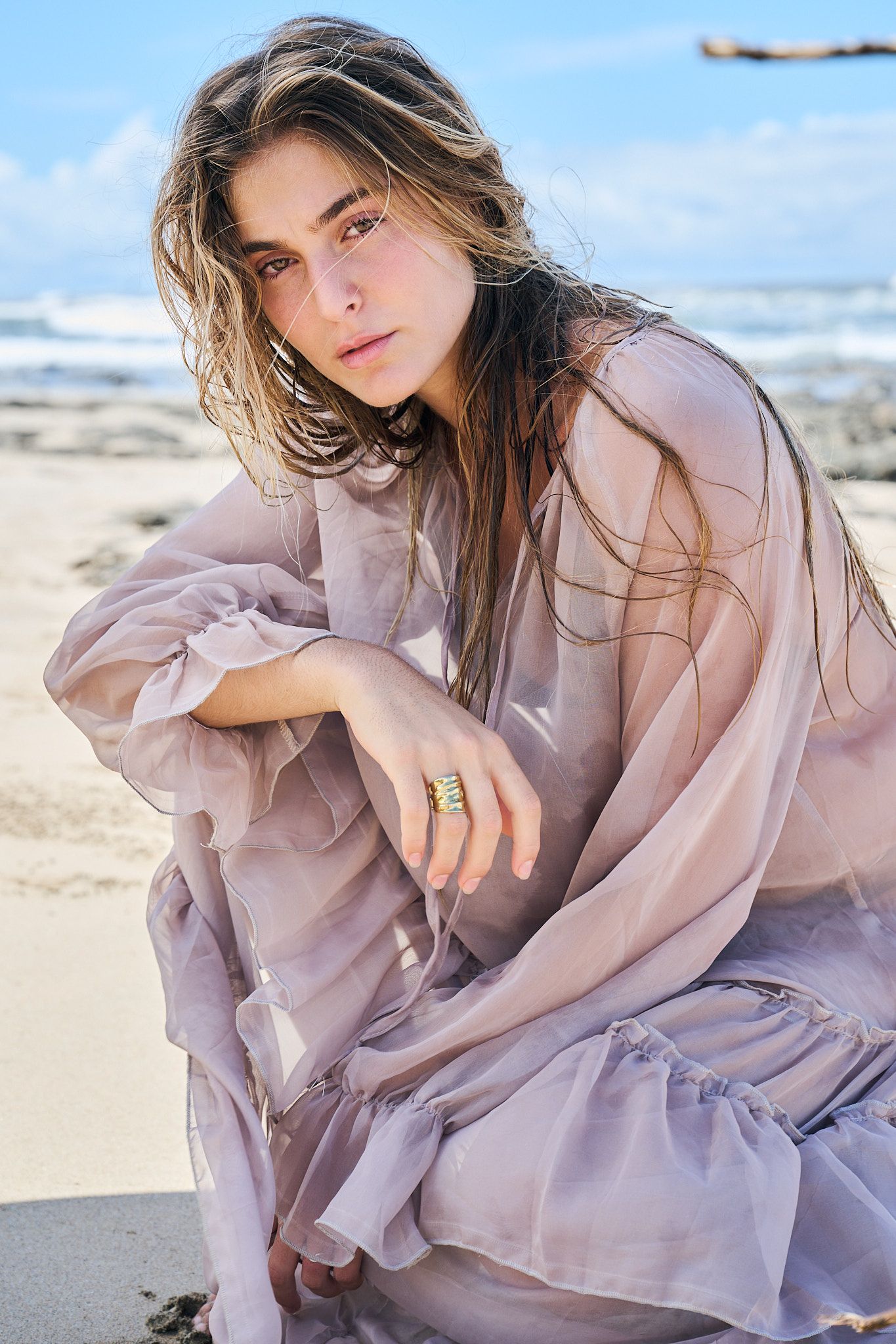 Person in flowy mauve dress poses artistically on sandy beach with soft natural lighting.