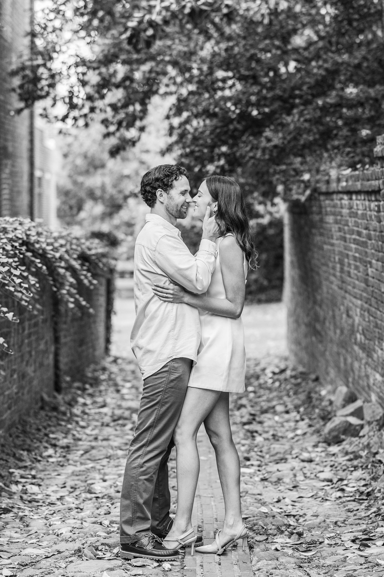 A couple shares a romantic moment in black and white while standing on a leaf-covered path next to a brick wall.