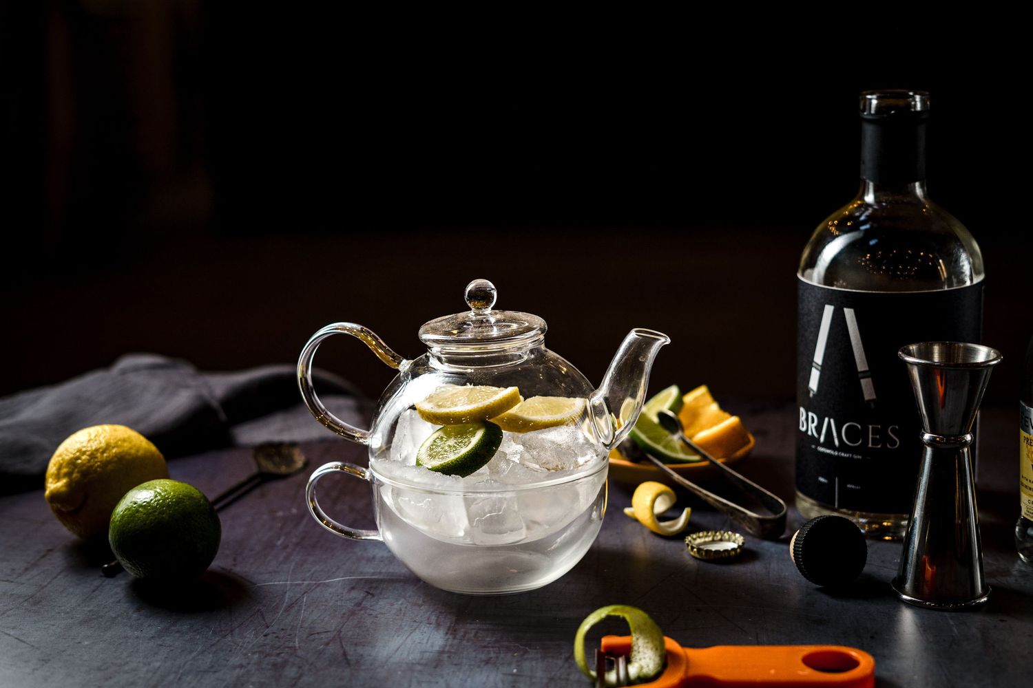 Brand photography of a cocktail preparation scene with citrus, ice, glass teapot and spirits bottle