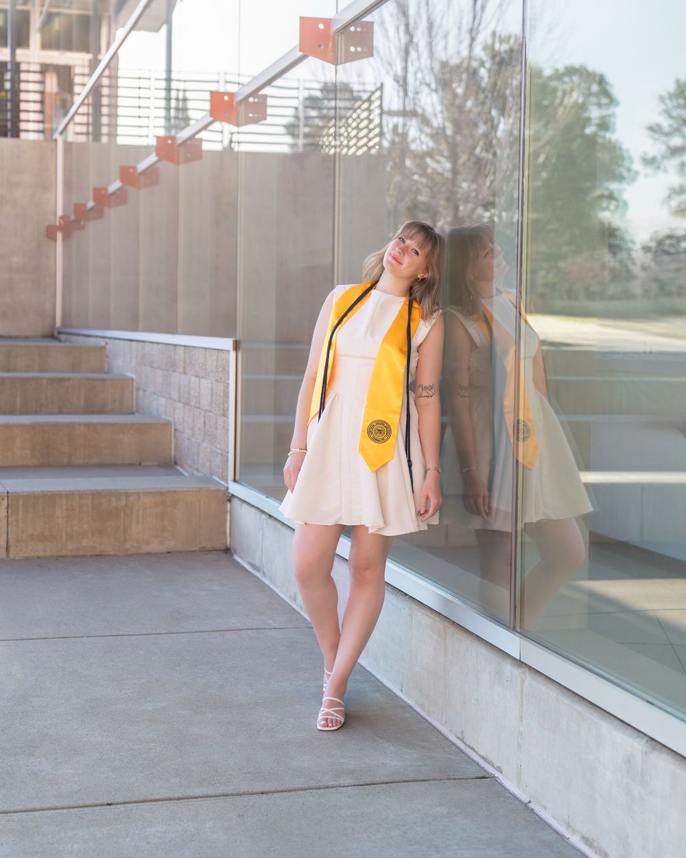 Graduate poses against glass railing during modern architectural graduation photo session.