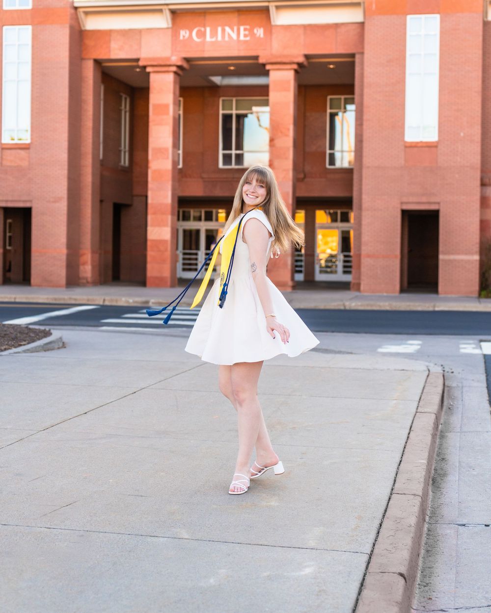 Graduate in white dress poses in front of red brick university building with diploma.