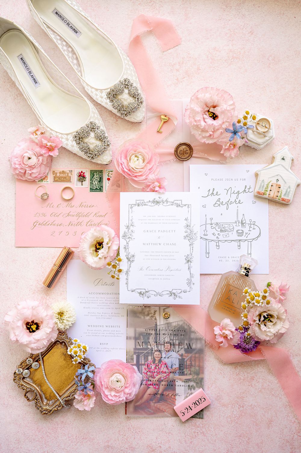 Wedding invitation flatlay with pink flowers, vintage stamps, and elegant white shoes on pink background.