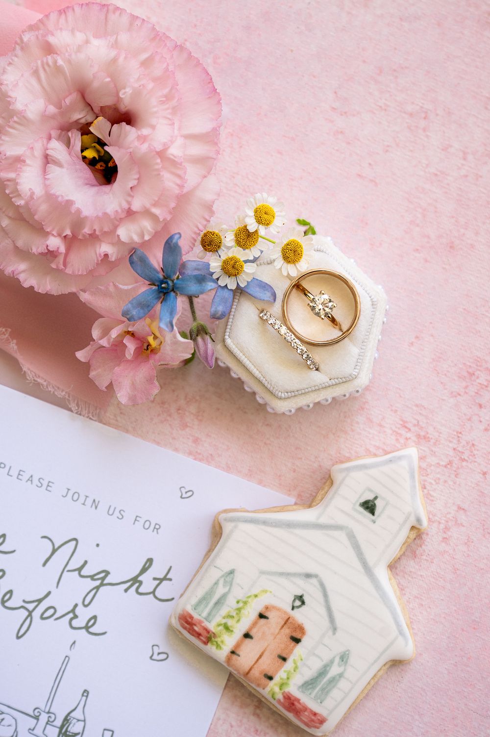Delicate wedding details including ring box, flowers, and invitation cards arranged on pink background.