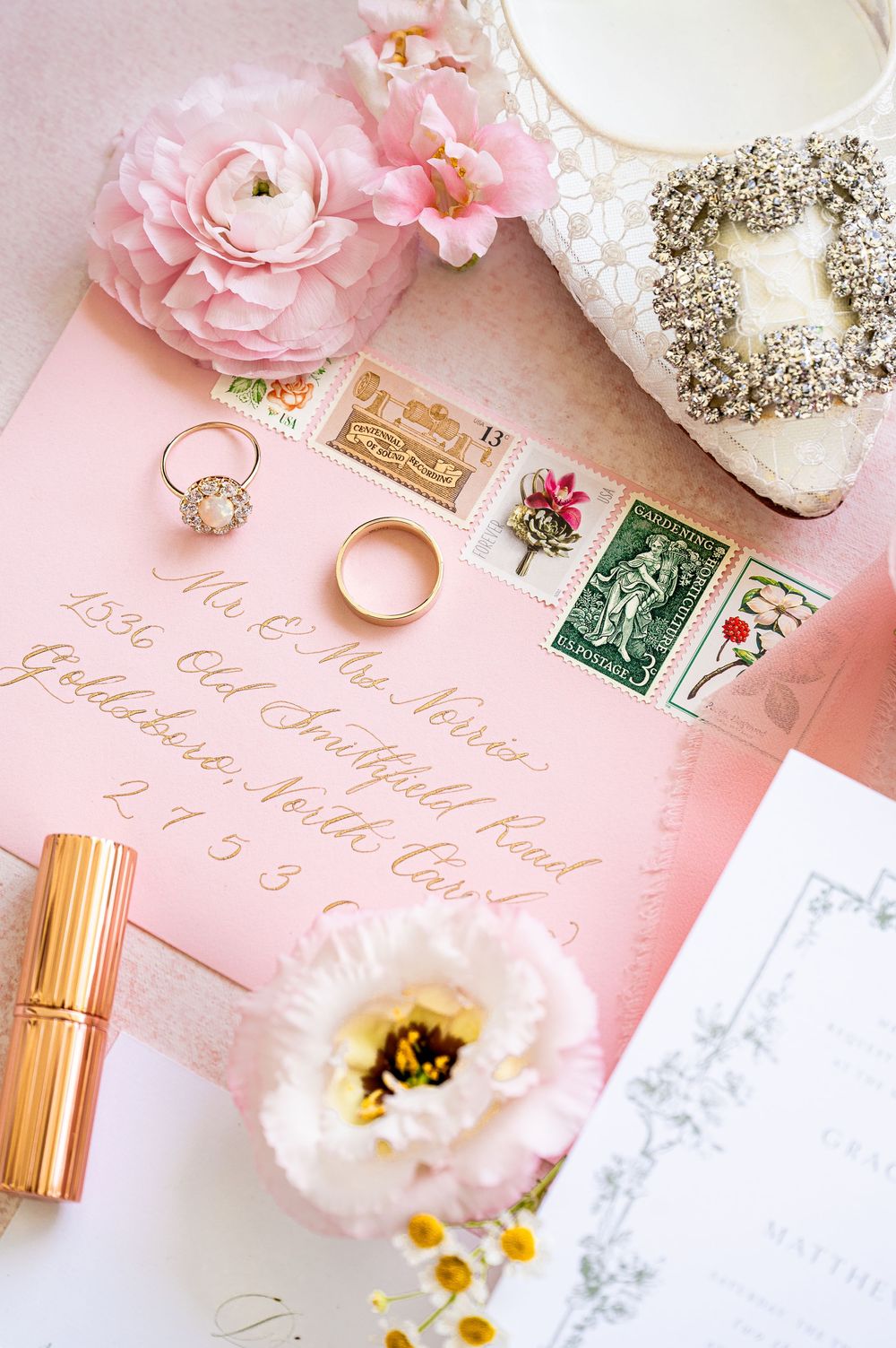 Wedding stationery and rings styled with pink flowers and vintage postage stamps on blush background.