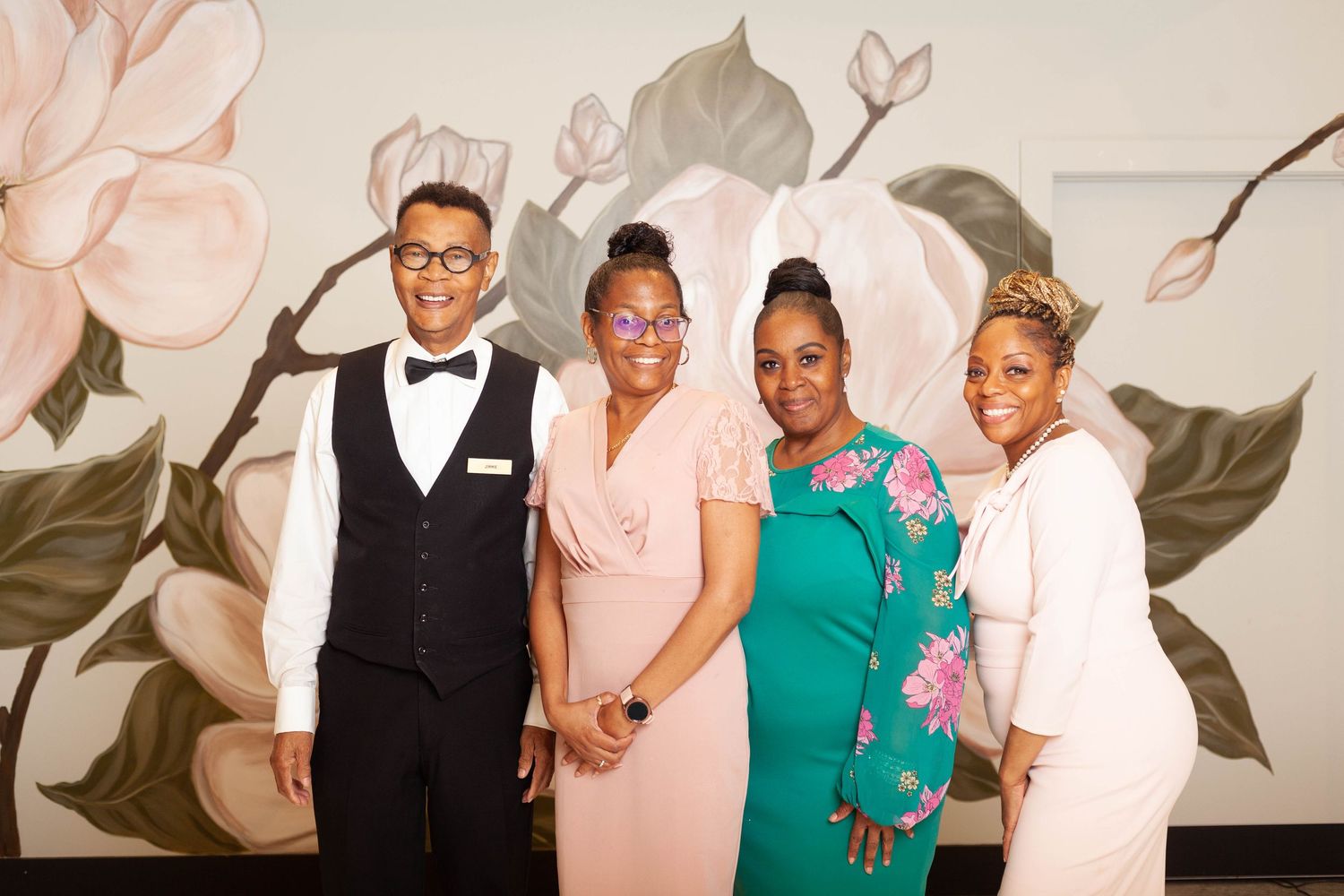 Four people in formal attire pose together against a floral wall backdrop at an elegant event.