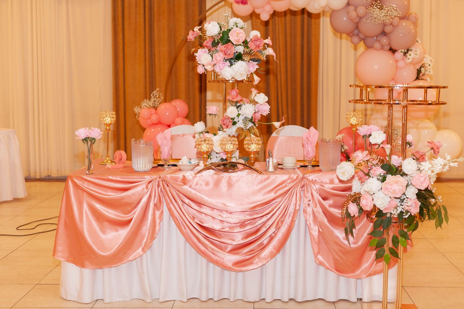 Elegant wedding reception decor featuring coral pink drapes, floral arrangements, and white tablecloths in a grand ballroom.