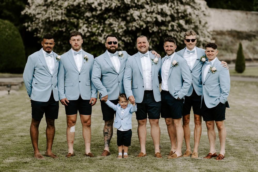 Wedding party wearing matching light blue blazers and navy shorts pose together outdoors with a small child in center.
