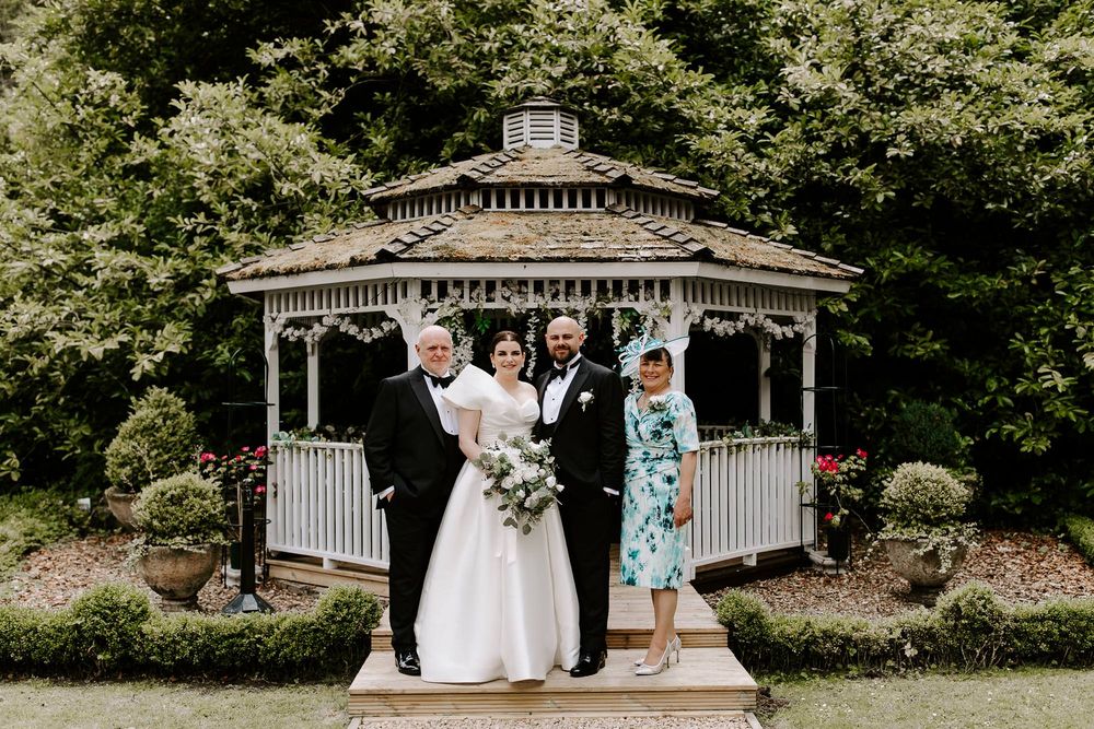 Wedding party gathers for outdoor photos at a gazebo surrounded by lush greenery.