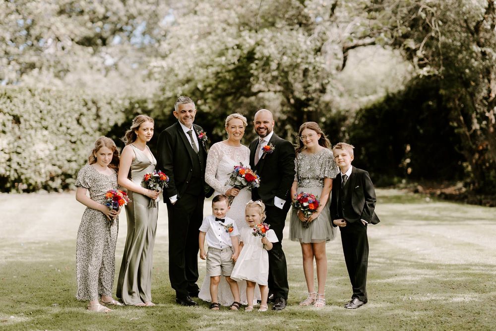 A wedding party group poses together outdoors on a sunny day in a garden setting.