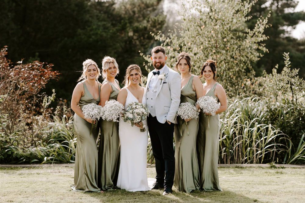 Wedding party poses in sage green and white attire against a natural garden backdrop.