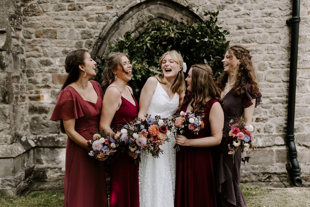 Wedding party laughs together while standing against a rustic stone wall backdrop.
