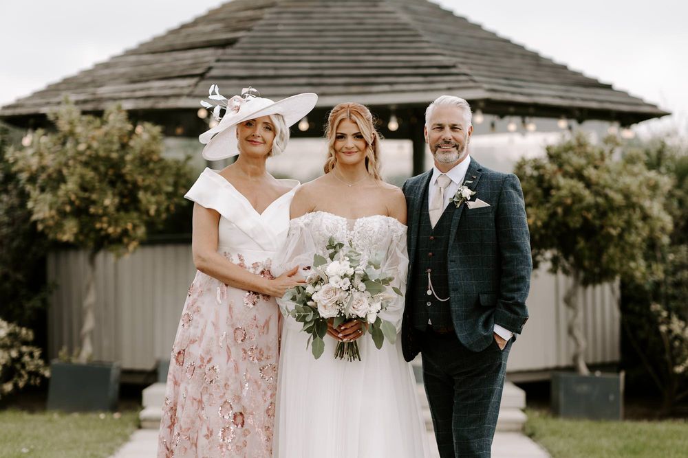Three people in formal wedding attire pose together outdoors with a white gazebo in the background.