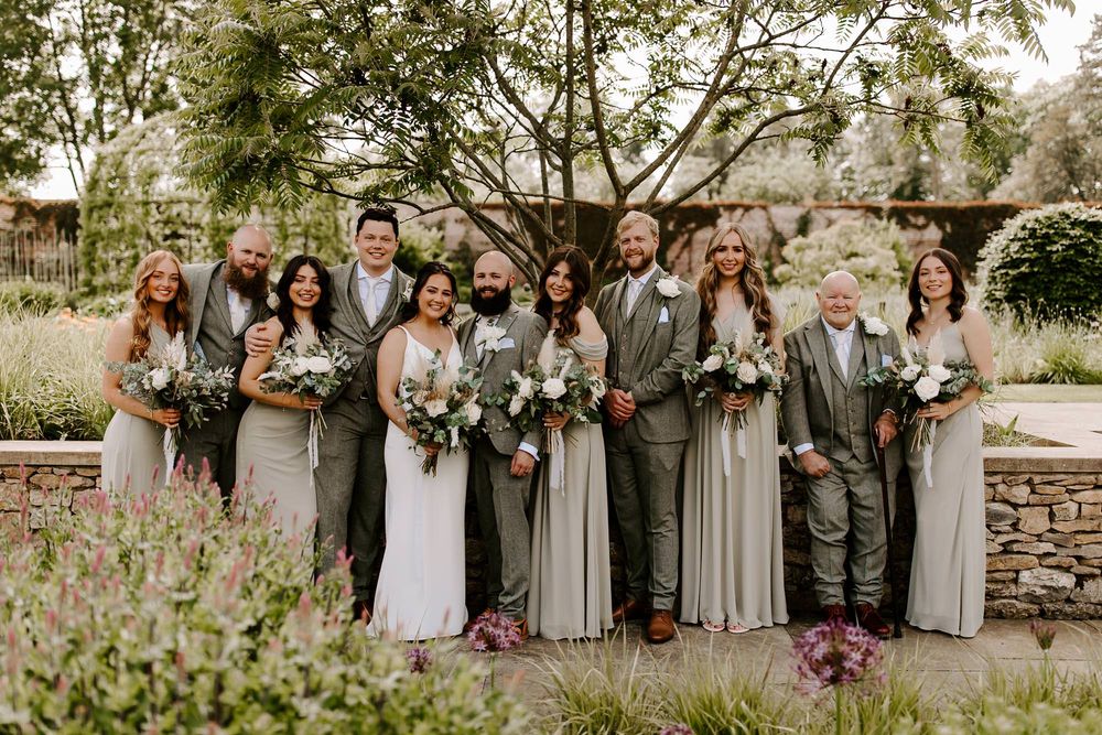 Wedding party in gray and white attire poses together in a garden setting with floral bouquets.