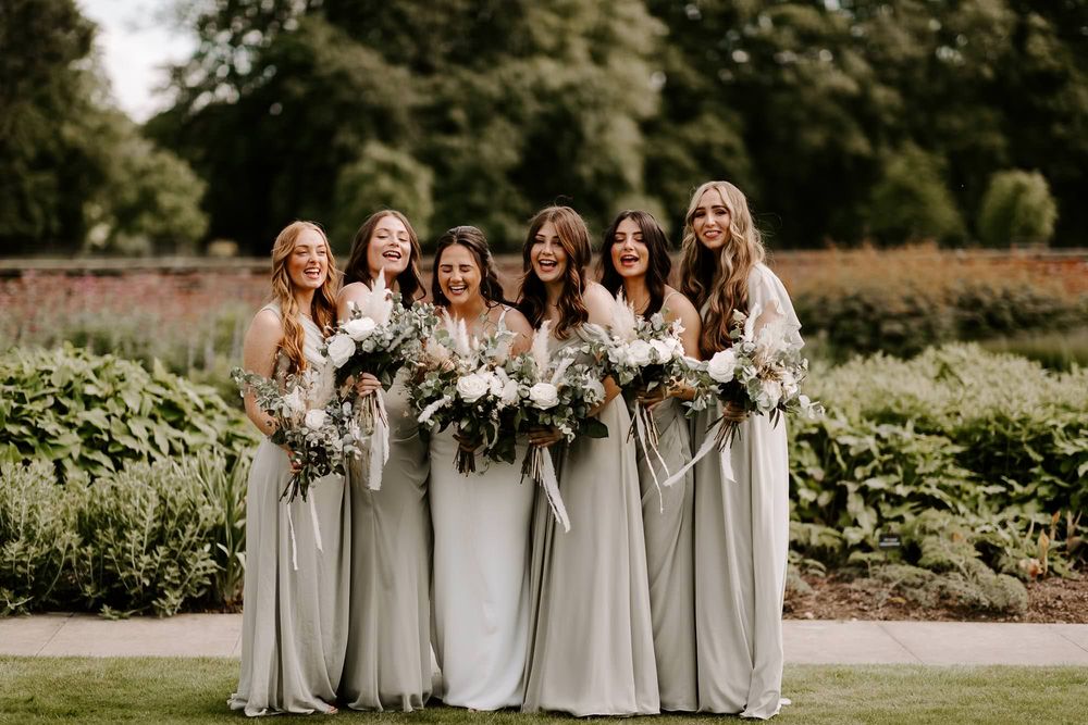 A wedding party in light gray dresses stands outdoors holding elegant white and green floral bouquets.