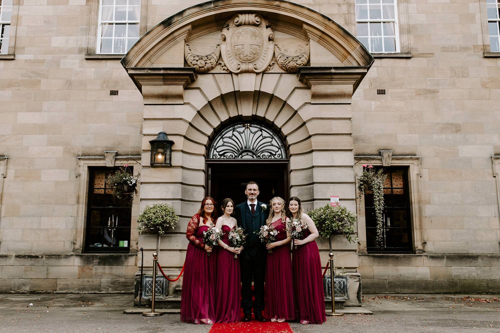 A historic stone building with elegant arched windows and ornate architectural details serves as backdrop for a wedding party.