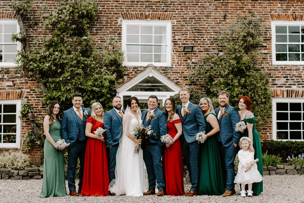 Wedding party in formal attire poses in line against rustic brick building exterior with white windows.