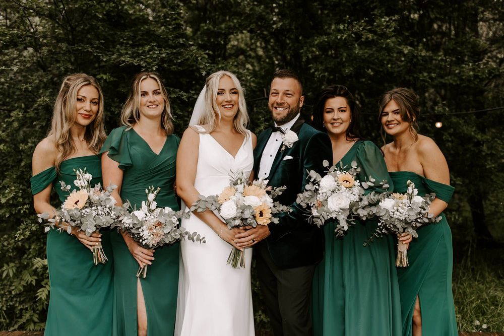 Wedding party members in emerald green dresses and formal attire pose with floral bouquets in an outdoor garden setting.
