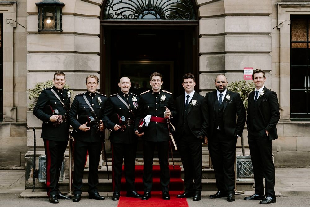 Wedding party in formal black attire poses on stone building steps with a red carpet.