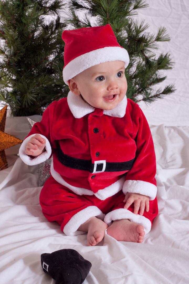 Baby boy with great big smile in Santa outfit with Christmas tree behind him.
