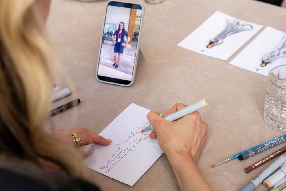 Close-up view of hands sketching on paper with various reference materials and drawings spread across a wooden desk.