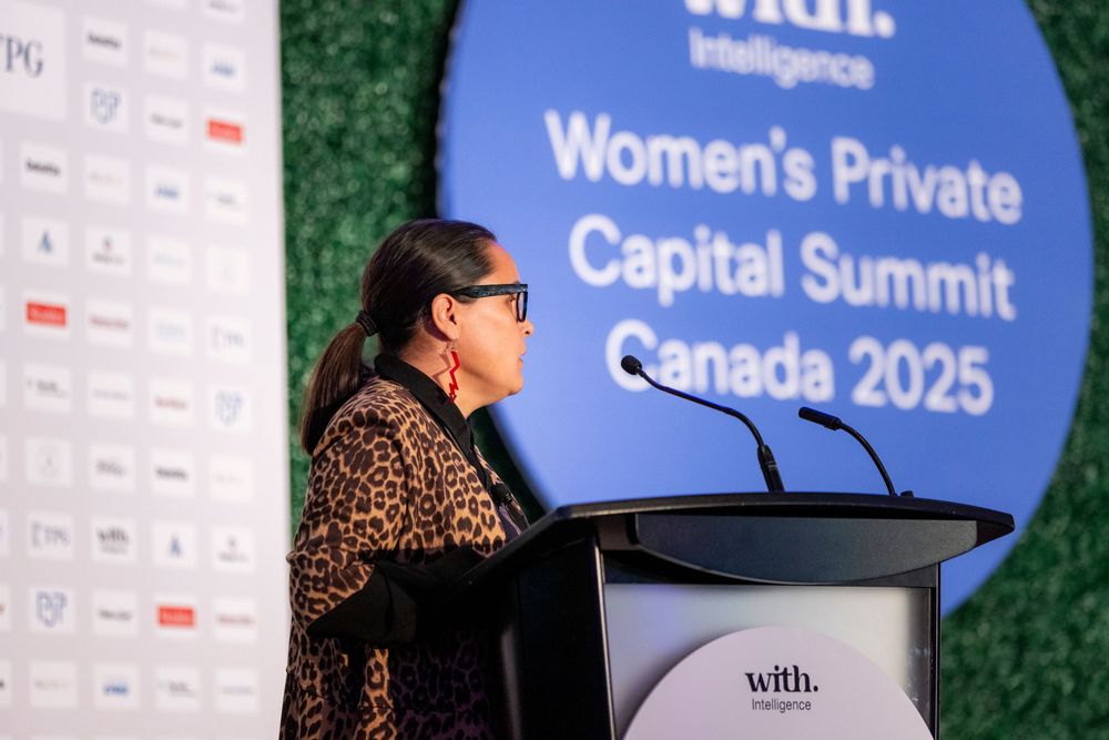 Speaker presenting at Women's Private Capital Summit with blue branded backdrop and sponsor wall.