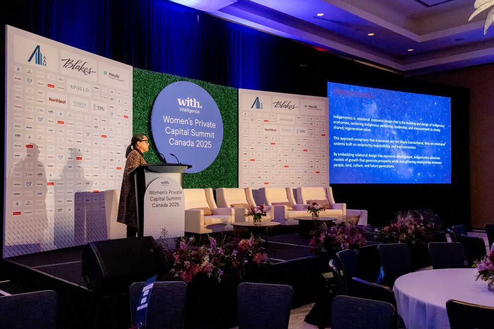 Conference speaker presenting at a business summit with corporate sponsor logos and blue lighting on stage.