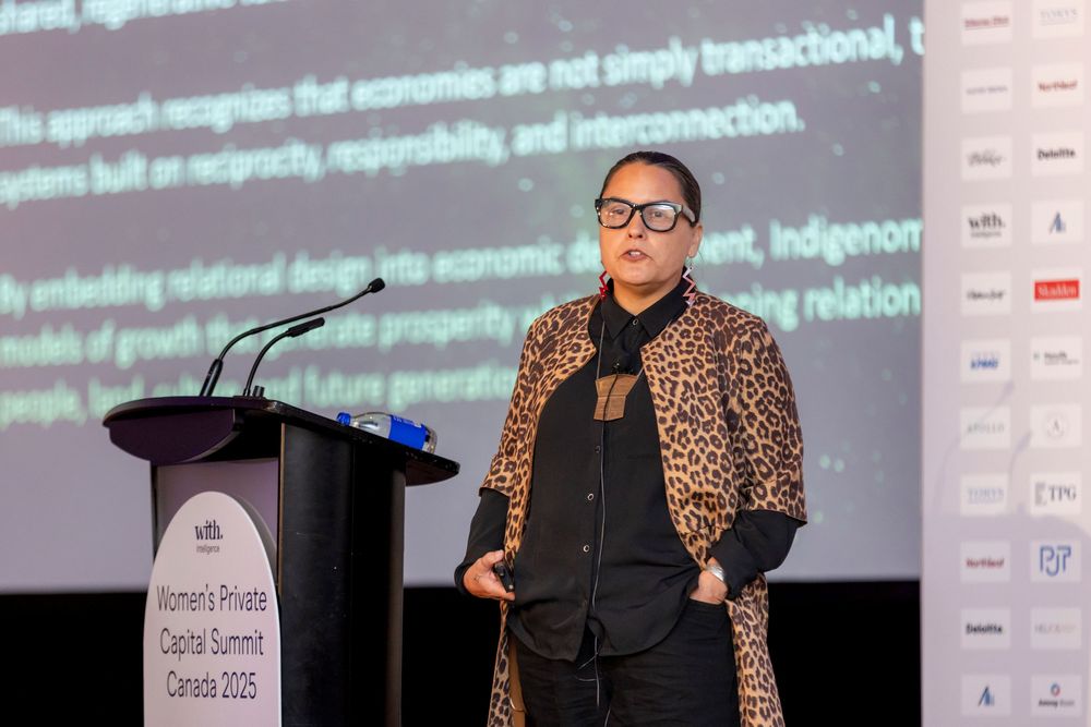 Conference presenter in leopard print jacket speaking at podium with presentation slides visible.