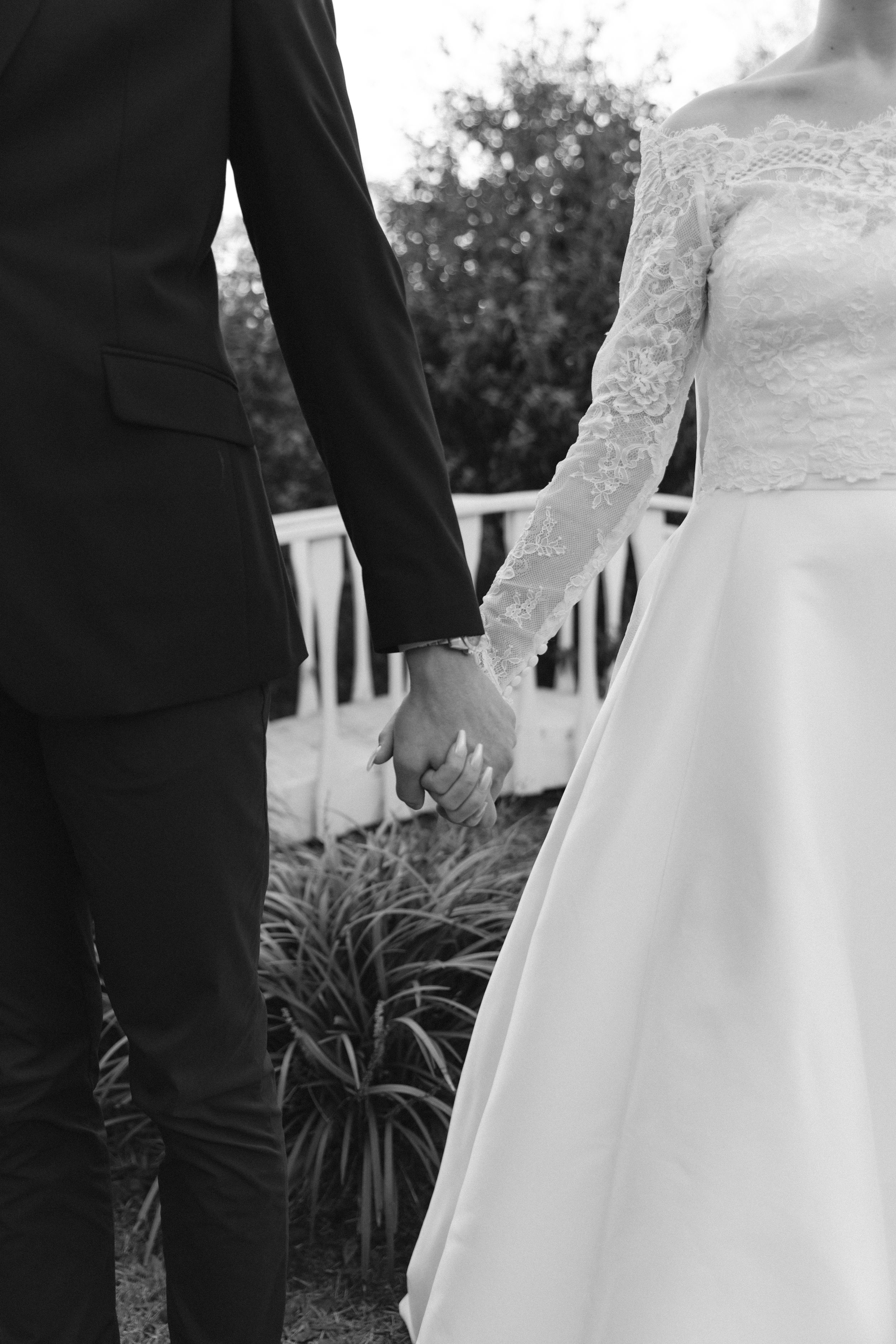 Black and white wedding photo showing couple holding hands with bride in lace dress and groom in dark suit.