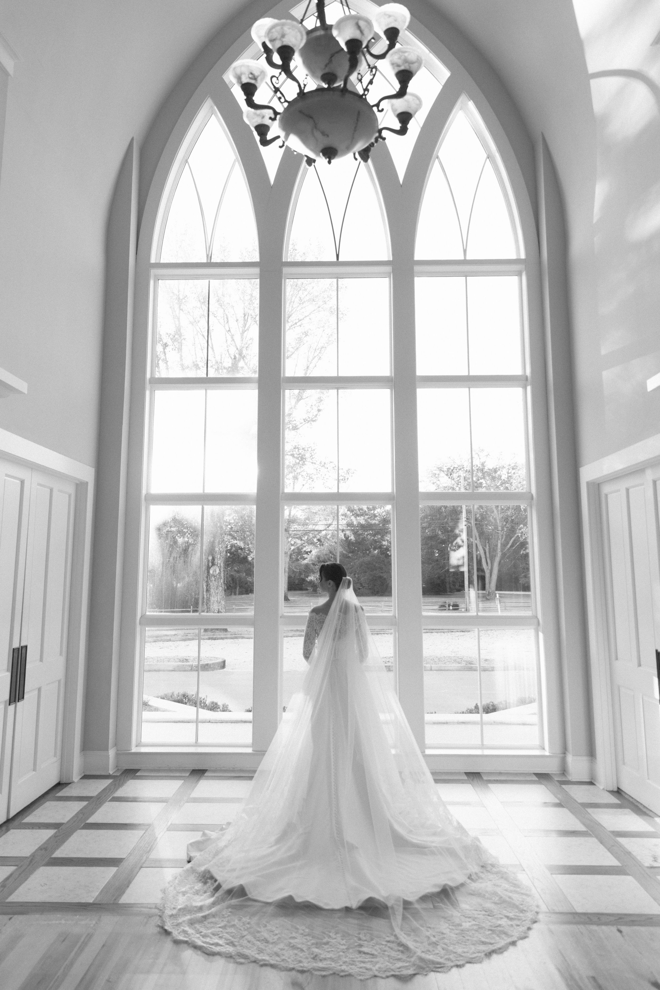 A wedding dress silhouette stands gracefully in front of arched windows in an elegant white chapel room.