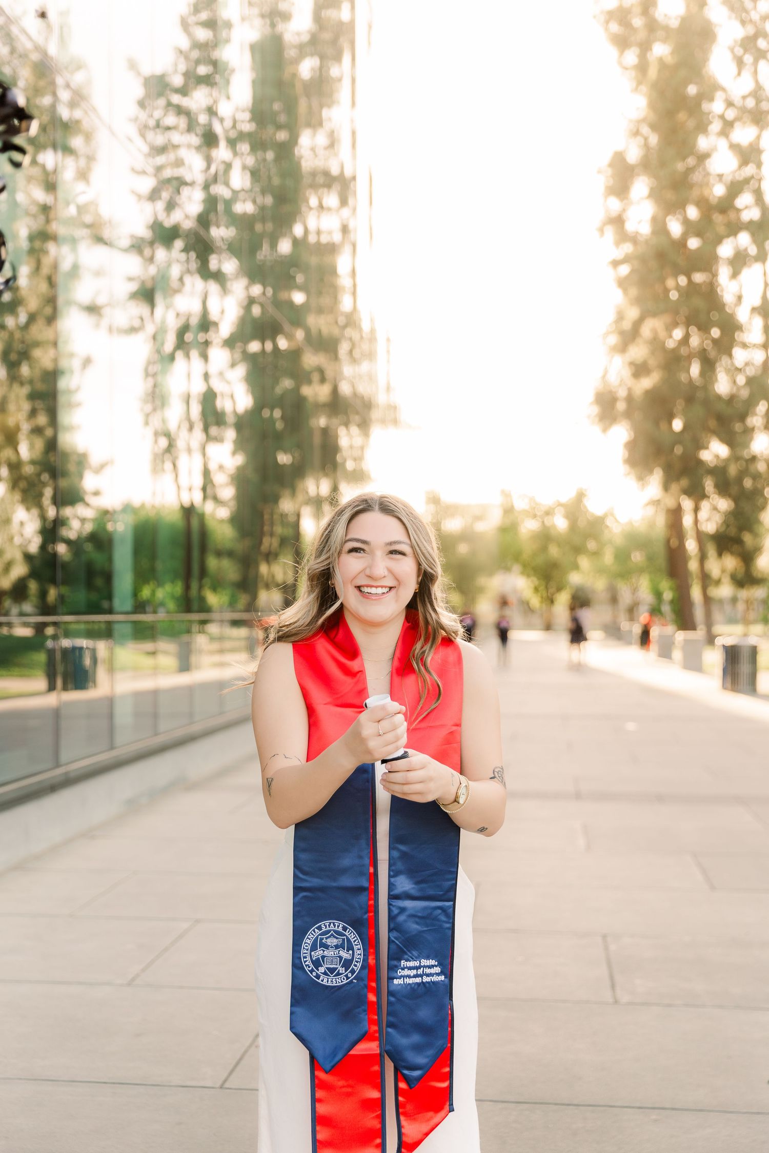 Graduate wearing a red and navy blue graduation stole poses outdoors with soft bokeh lighting in the background.