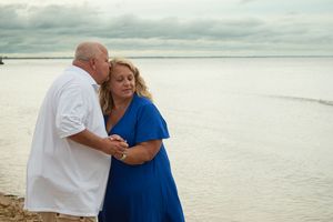 A couple in white and blue clothing share an intimate moment on a peaceful beach under cloudy skies.