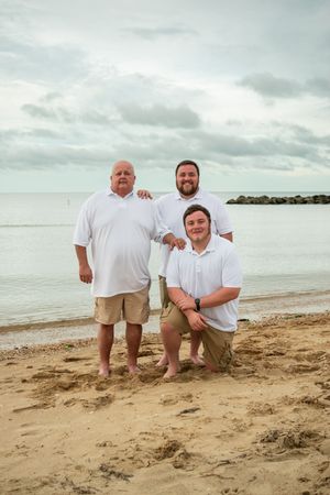 Group in white shirts poses casually on a sandy beach with ocean and cloudy sky in the background.
