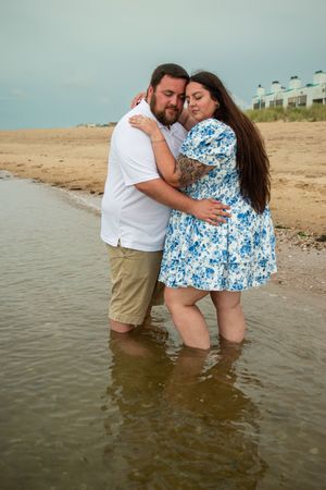 A romantic couple shares intimate moments while standing in shallow water at the beach wearing white and blue floral attire.
