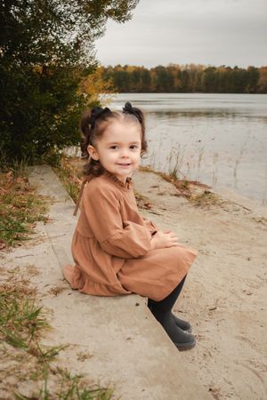 Small child plays and poses on a sandy beach shore near a lake during autumn.