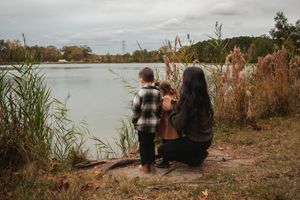 Two people sit by a serene lake surrounded by tall grasses and autumn foliage at sunset.