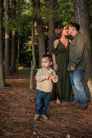 Family portrait series in a pine forest with toddler in khaki shirt and jeans standing in foreground.