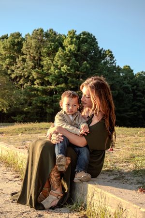 A tender outdoor portrait of a parent and child sitting together on a stone wall at sunset with pine trees in the background.