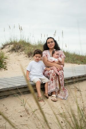 A mother and child sit together on a wooden boardwalk along sandy beach dunes on a cloudy day.