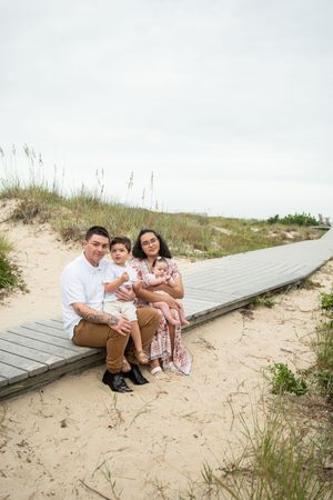 A family sits together on a wooden boardwalk path through sand dunes at the beach on a sunny day.
