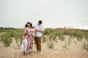 Family wearing coordinated white and brown outfits poses together on a sandy beach with dune grass in background.