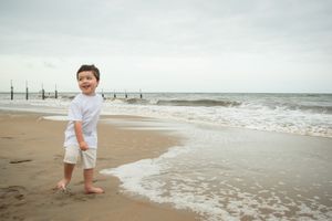 A small child stands barefoot on a sandy beach as waves roll in on an overcast day.