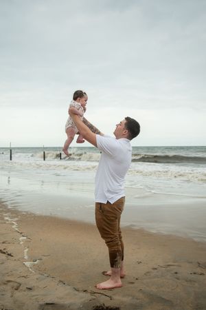 Parent lifts and plays with child on sandy beach with ocean waves in background.