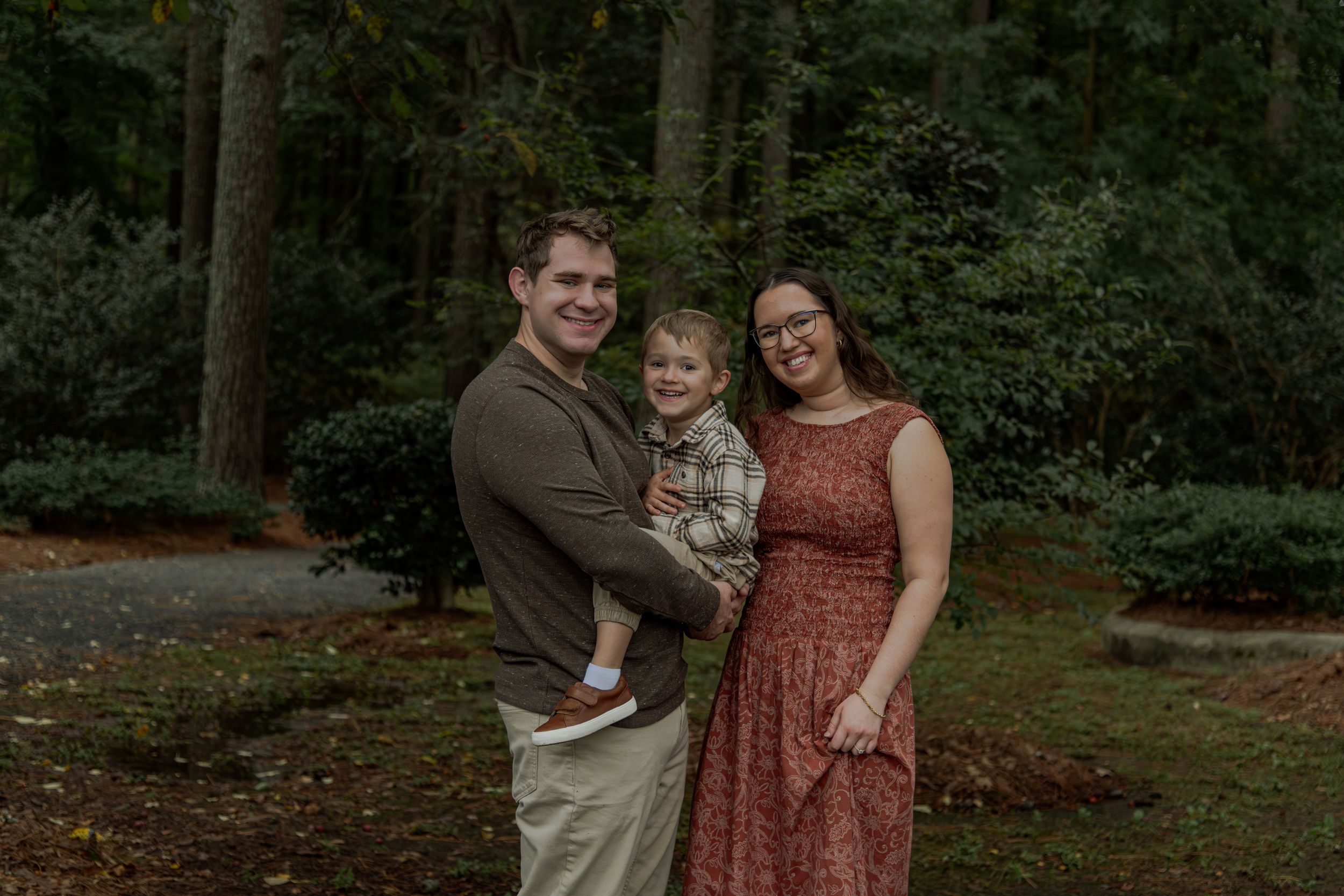 A couple stands together in a wooded outdoor setting wearing coordinated fall-toned outfits.