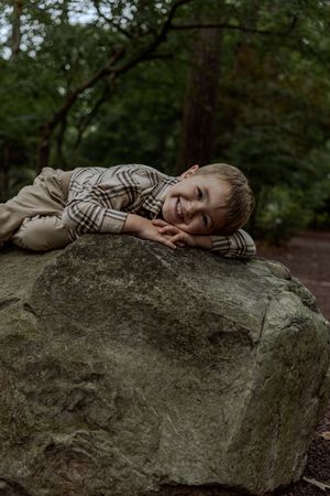 A young child lies peacefully asleep on a large gray boulder in a wooded forest setting.